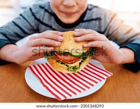 Child, hands and eating burger on table for delicious lunch, meal or food with healthy vegetables at home. Hand of hungry little boy holding beef hamburger for fresh dinner, nutrition or vitamins