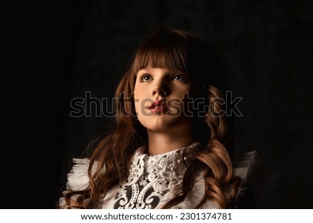 Face of stylish cover little girl in doll image isolated in shadow, frozen look looking away. Charming kid 9-10 year old with curly hair at black background. Fashion style concept. Copy ad text space