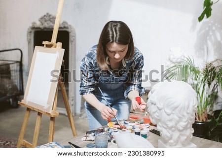 Interior of artist's workshop. Creative person concept. Easel. Woman paints a picture. The artist chooses colors for the painting. She is wearing denim overalls and plaid shirt. Mock up.