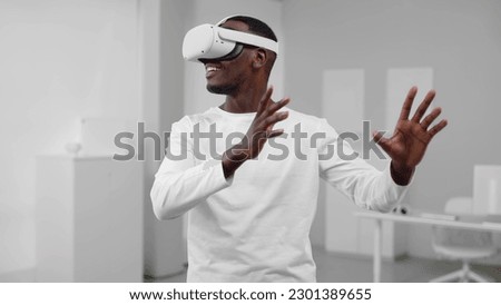 Portrait of African-American man in white clothes use augmented reality headset standing in room with white walls and furniture. 