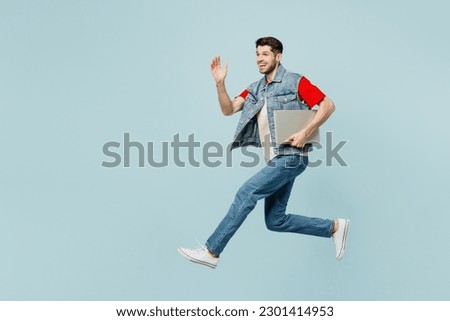 Full body side view young IT man wear denim vest red t-shirt casual clothes jump high hold closed laptop pc computer isolated on plain pastel light blue background studio portrait. Lifestyle concept