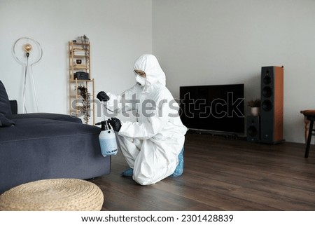 Cleaning worker in protective suit disinfecting furniture using sprayer with detergent during work in apartment Royalty-Free Stock Photo #2301428839