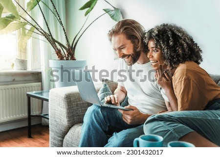 Multiracial young couple watching computer laptop sitting on the sofa at home - Happy diverse husband and wife using pc online services - Technology life style concept  Royalty-Free Stock Photo #2301432017