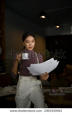 Portrait of a professional millennial Asian female tailor or fashion designer focuses on checking her model sketches while sipping coffee in a studio.
