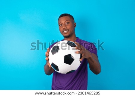 Portrait of smiling young man wearing purple T-shirt holding a ball over blue studio background looking at camera and gesturing finger frame. Creativity and photography concept.