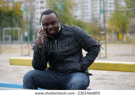 African american man sits at sport ground, speaks on smartphone and smiles
