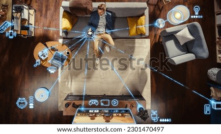 Top View Of Caucasian Man In the Loft Apartment Sitting Down on The Couch and Connecting Smartphone to Convenient Smart Home System. VFX Edit Visualizing Connected Devices. Laptop, TV, Speaker. Royalty-Free Stock Photo #2301470497