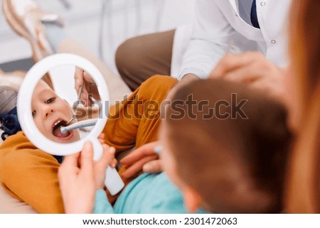 Dentist fixing tooth to a brave little boy sitting in dentist chair in mother's lap using dental drill and angled mirror Royalty-Free Stock Photo #2301472063
