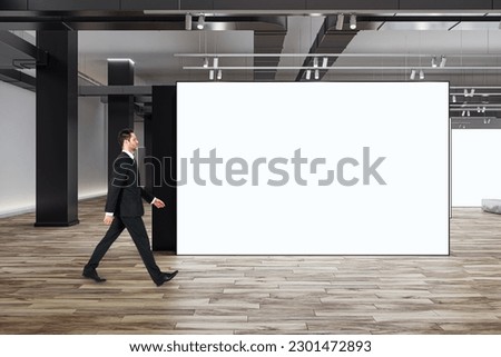 Marketing concept with businessman walking by blank white partition with space for advertising poster or picture frame in loft style gallery hall with wooden floor and grey wall background, mock up Royalty-Free Stock Photo #2301472893
