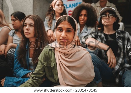 Muslim girl looking at the camera while sitting with a group of climate activists outside a building. Multicultural youth demonstrators protesting against global warming and climate change.