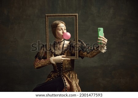 Portrait of pretty young girl, princess with bubble gum, holding picture frame and taking selfie with phone against dark green background. Concept of history, renaissance art, comparison of eras Royalty-Free Stock Photo #2301477593
