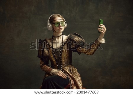 Portrait of young beautiful girl in vintage dress, trendy sunglasses and headphones, taking selfie with phone against dark green background. Concept of history, renaissance art, comparison of eras Royalty-Free Stock Photo #2301477609