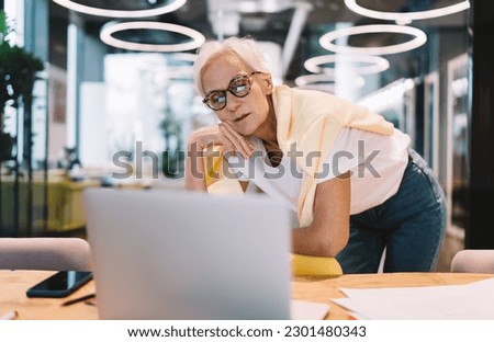 Unsure aged female businesswoman in eyeglasses standing at table with laptop and looking at screen while waiting for updates from project