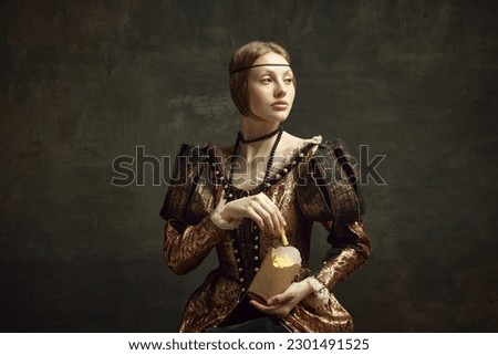 Portrait of young beautiful woman in vintage costume, eating fries against dark green background. Fast food lover. Concept of history, renaissance art, comparison of eras, health and modern food