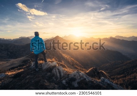 Man on stone on the hill and beautiful mountain valley in haze at colorful sunset in autumn. Dolomites, Italy. Guy, mountain ridges in fog, orange grass and trees, blue sky with sun in fall. Hiking	 Royalty-Free Stock Photo #2301546101