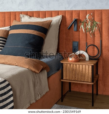 Minimalist composition of bedroom interior with orange bed, beige bedclothes, wooden night stand, vase with leaves, modern lamp on the wall, clock and personal accessories. Home decor. Template. 