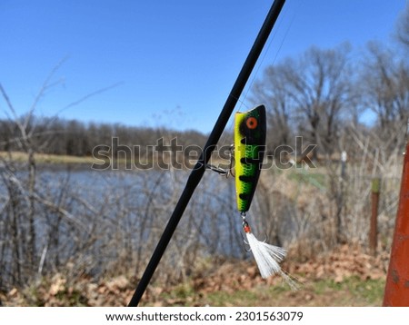 A close-up of a bass lure dangling from a fishing pole and a pond in the background calls to the fisherman. Rural Missouri, MO, United States, USA, US