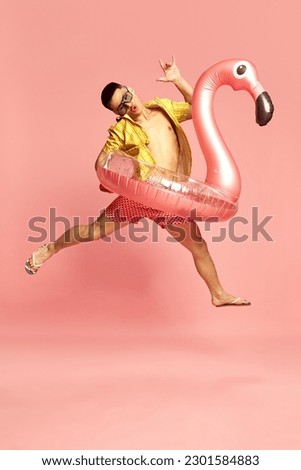 Pool party. Portrait with attractive young guy, man wearing beach clothes jumping with inflatable flamingo over pink studio background. Concept of human emotions, celebrating, vacation, summer Royalty-Free Stock Photo #2301584883