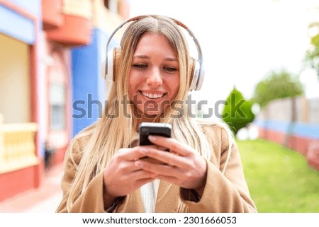 Young pretty blonde woman at outdoors listening music and looking to mobile