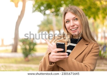 Young pretty blonde woman at outdoors using mobile phone and pointing back