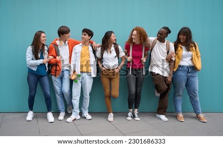 Multiracial group of cheerful erasmus students enjoying free time. Young friends together leaning against a blue wall and looking at each other smiling. Generation z and youth relationships. Royalty-Free Stock Photo #2301686333