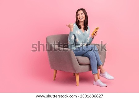 Full size photo of young girl brown hair sit armchair waiting favorite store point finger empty space phone isolated on pink background
