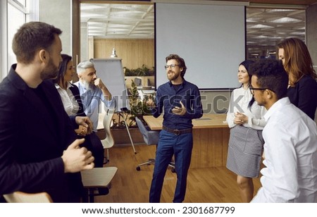 Young man business coach trainer talking to group of people in office standing around him and listening. Office workers, entrepreneurs on corporative training, team building seminar or master class. Royalty-Free Stock Photo #2301687799