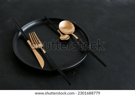 Empty black plate and black gold cutlery set top view with copy space. Black kitchen utensils set on black textured background. Abstract geometric food concept. Toned