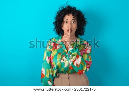 young arab woman wearing colorful shirt over blue background makes silence gesture, keeps finger over lips. Silence and secret concept.