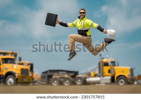 Middle aged builder excited jump on site construction. Excited builder construction worker in a safety helmet jumping in front of the trucks. Excited crazy builder man in helmet jump outdoor. Royalty-Free Stock Photo #2301757815