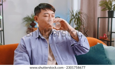 Thirsty asian man holding glass of natural aqua make sips drinking still water preventing dehydration sits at home living room. Chinese Guy with good life habits, healthy slimming, weight loss concept Royalty-Free Stock Photo #2301758677