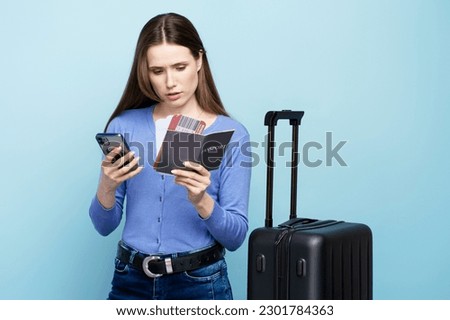 Young woman tourist using smartphone, checking mobile app, online registration of flight, holding boarding pass, posing with suitcase over blue background. Travel tourism journey people technology  Royalty-Free Stock Photo #2301784363