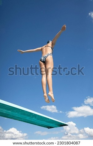 View from below. Fit woman jumping high off a diving board into the swimming pool. Abstract image with blue sky and clouds in the background. Lots of copy space. Royalty-Free Stock Photo #2301804087