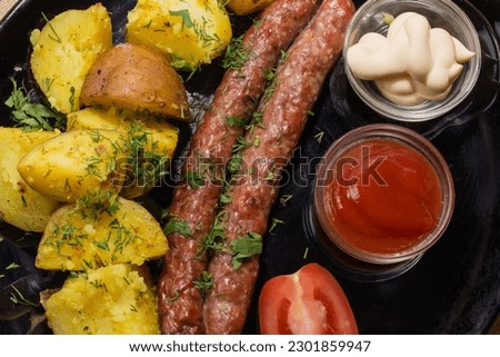 Grilled thin long sausages with baked potatoes and different sauces on the black dish, top view close-up
