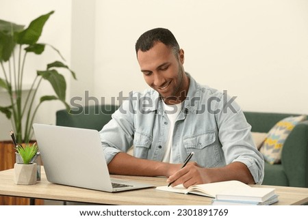 African American man writing in notepad near laptop at wooden table in room