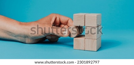 closeup of a man putting a cylindrical building block in a stack of rectangular building blocks, on a blue background, in a panoramic format to use as web banner or header Royalty-Free Stock Photo #2301892797