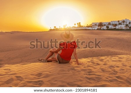 A tourist woman with a sunhat in Maspalomas Dunes of Gran Canaria sunset. She either listens to distant ocean waves or watches sand dunes shift in the breeze while closing her eyes. Royalty-Free Stock Photo #2301894181