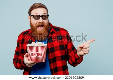 Young fun ginger man in 3d glasses wear casual clothes watch movie film hold bucket of popcorn in cinema point index finger aside on area isolated on plain pastel blue cyan background studio portrait