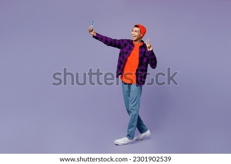Full body young man of African American ethnicity wear casual shirt orange hat doing selfie shot on mobile cell phone post photo on social network show v-sign isolated on plain purple color background