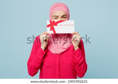 Young arabian muslim woman wear pink abaya hijab hold cover mouth with gift certificate coupon voucher card for store isolated on plain blue cyan background studio. People uae islam religious concept