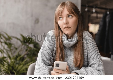 Confused puzzled blonde bang woman in casual clothes, sits in cafe, holds a smartphone in her hand, looks questioningly, rolling eyes. Girl getting surprising bad news, annoyed woman. Royalty-Free Stock Photo #2301924159
