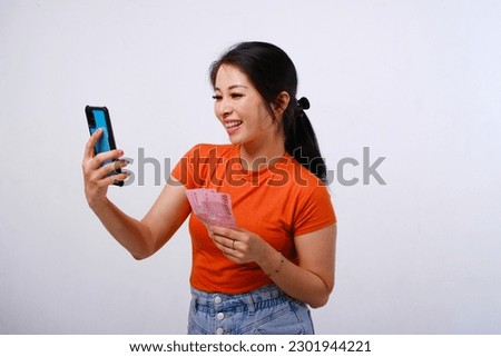 Excited and happy Asian woman wearing casual shirt holding Indonesian money and phone isolated on white background