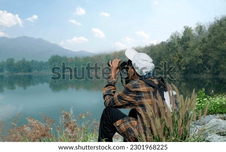 Asian boy in plaid shirt wears white cap, holding binoculars, sitting on reservoir ridge during summer vacation and birdwatching activity, soft and selective focus, nature study and hobby concept. Royalty-Free Stock Photo #2301968225