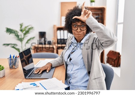 Black woman with curly hair wearing call center agent headset at the office making fun of people with fingers on forehead doing loser gesture mocking and insulting. 