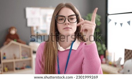 Young blonde woman working as teacher standing serious expression at kindergarten