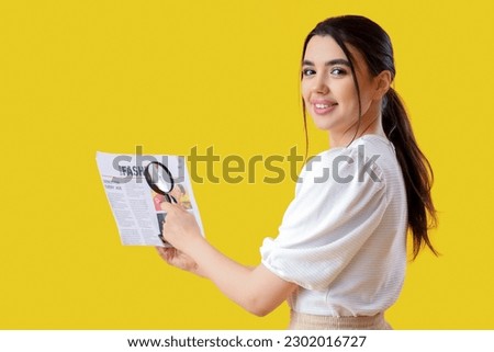 Young woman with magnifier reading newspaper on yellow background