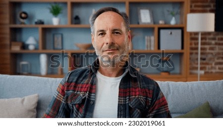 Portrait of likable satisfied positive modern bearded man in checkered shirt sitting on comfortable sofa in living-room in front of camera with sincerely smile Royalty-Free Stock Photo #2302019061