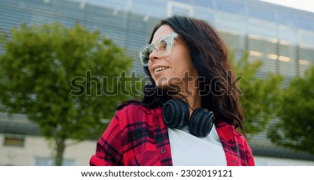 Splendid positive happy modern stylish young woman in casual clothes in glasses with headphones looking around on the background of beautiful urban building