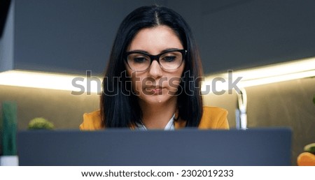 Likable concentrated confident busy dark-haired woman in glasses working on computer in kitchen background and making break to drink water from glass Royalty-Free Stock Photo #2302019233