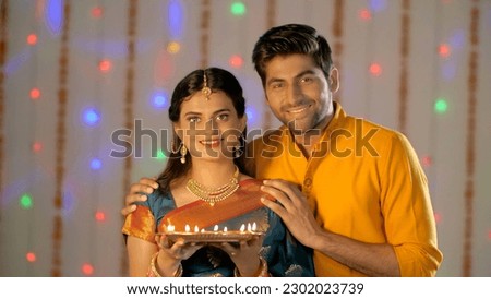 A newly married couple celebrating Diwali with brightly lighted Diyas. Medium shot of an Indian husband-wife in traditional clothes standing with a plate full of oil lamps Royalty-Free Stock Photo #2302023739